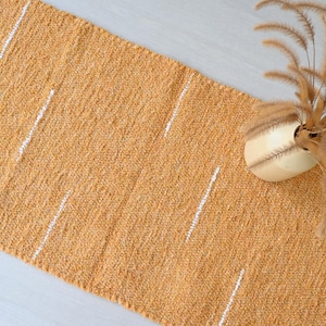 Puede incluir: Una alfombra rectangular texturizada en un tono amarillo dorado cálido, acentuada con rayas blancas verticales. La alfombra está hecha de un material tejido. Un jarrón crema con hierba ornamental seca se encuentra sobre la alfombra.