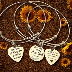 Op de afbeelding: Drie zilverkleurige, verstelbare armbanden met hartvormige bedels. Elke bedel heeft een inspirerende boodschap: "Met God zijn alle dingen mogelijk", "Dankbaar, Dankbaar, Gezegend" en "Vertrouw op de Heer met heel je hart". De armbanden worden gepresenteerd met zonnebloemen.