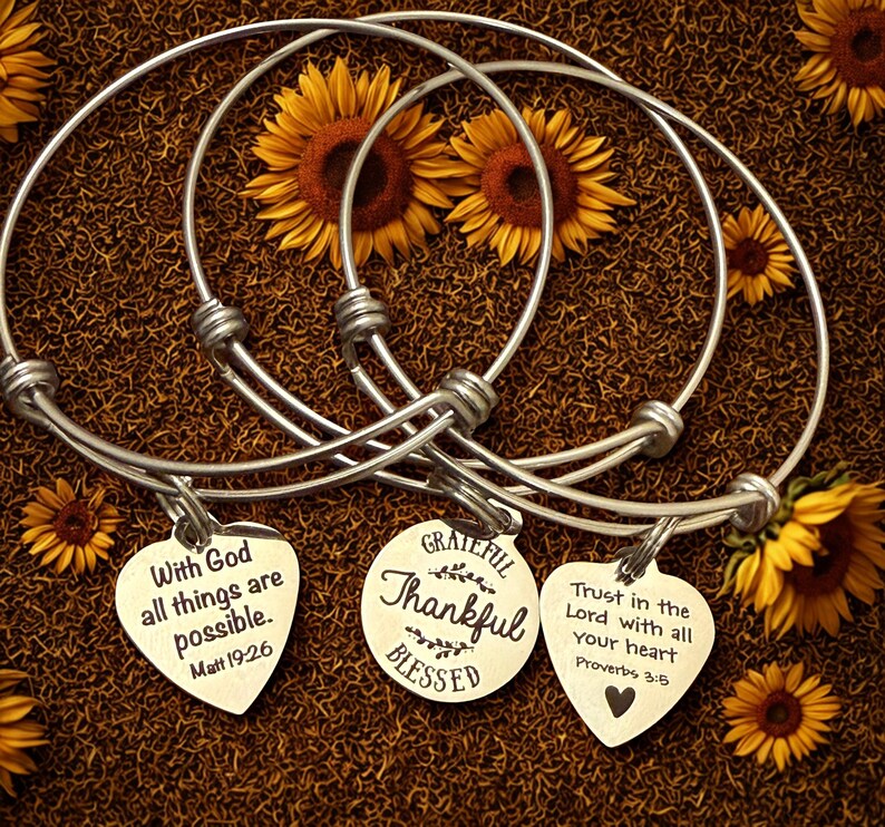 Op de afbeelding: Drie zilverkleurige, verstelbare armbanden met hartvormige bedels. Elke bedel heeft een inspirerende boodschap: "Met God zijn alle dingen mogelijk", "Dankbaar, Dankbaar, Gezegend" en "Vertrouw op de Heer met heel je hart". De armbanden worden gepresenteerd met zonnebloemen.