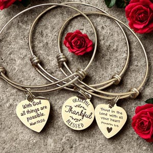 May include: Three silver-toned adjustable bangles with heart and round charms. The charms have inspirational quotes: "With God all things are possible," "Grateful, Thankful, Blessed," and "Trust in the Lord with all your heart." Red roses are in the background.