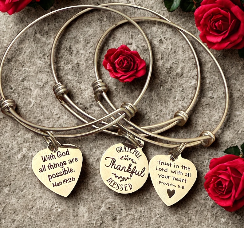Op de afbeelding: Drie zilverkleurige verstelbare armbanden met hart- en ronde bedels. De bedels hebben inspirerende citaten: "Met God zijn alle dingen mogelijk", "Dankbaar, Dankbaar, Gezegend" en "Vertrouw op de Heer met heel je hart". Rode rozen op de achtergrond.