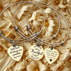 Op de afbeelding: Zilverkleurige armbanden met hartvormige en ronde bedels. De bedels hebben inspirerende citaten: "Met God zijn alle dingen mogelijk", "Dankbaar, Dankbaar, Gezegend" en "Vertrouw op de Heer met heel je hart."