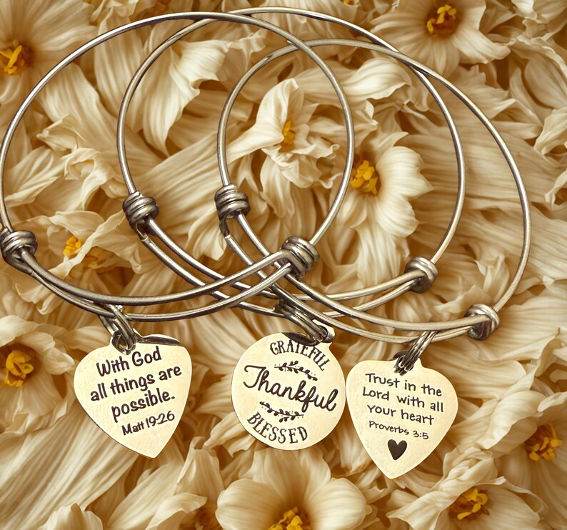 Op de afbeelding: Zilverkleurige armbanden met hartvormige en ronde bedels. De bedels hebben inspirerende citaten: "Met God zijn alle dingen mogelijk", "Dankbaar, Dankbaar, Gezegend" en "Vertrouw op de Heer met heel je hart."