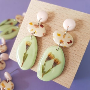May include: A pair of dangle earrings with a green resin base and pressed flowers. The top portion of the earrings feature a white resin base with brown, yellow, and pink flecks.