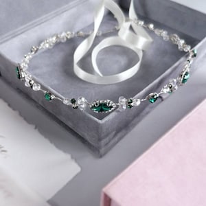 Puede incluir: Una corona de pelo de novia plateada y verde esmeralda con cristales transparentes, expuesta en una caja de terciopelo gris con una cinta de satén blanca.
