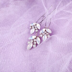 May include: Three silver hair pins with white crystal embellishments in the shape of leaves. The pins are on a light purple fabric background.
