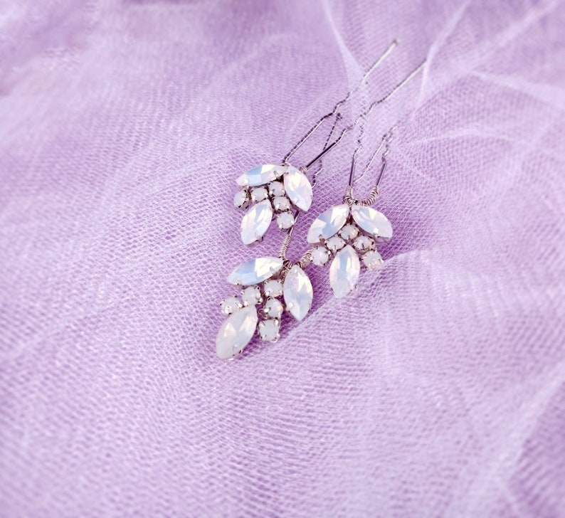 May include: Three silver hair pins with white crystal embellishments in the shape of leaves. The pins are on a light purple fabric background.