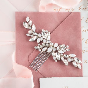 May include: A silver hair comb with a delicate design featuring a cluster of clear and iridescent crystals. The comb is set against a blush pink background.