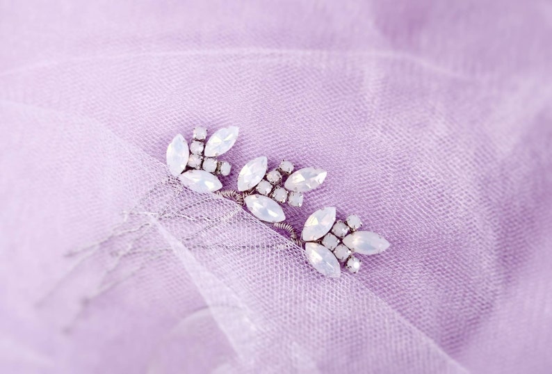 May include: Three silver hair combs with white opal-like stones arranged in a leaf design. The combs are on a light purple background.