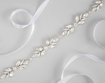 May include: A decorative belt featuring a repeating leaf-like pattern composed of clear, faceted crystals and small white pearls. The crystal embellishment is attached to a long, smooth white satin ribbon that extends from both ends of the piece.