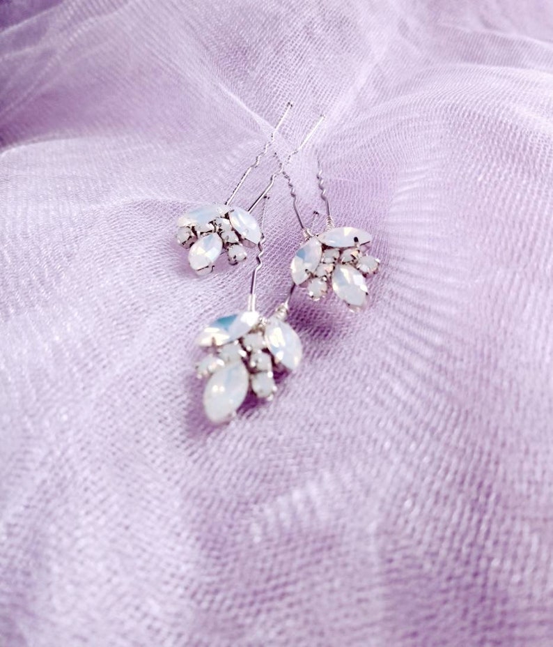 May include: Three silver hair pins with clusters of clear iridescent rhinestones. The pins are on a light purple fabric background.
