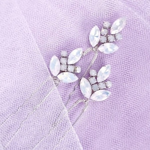 May include: Three silver hair clips with white crystal embellishments. The clips are arranged in a cluster on a light purple background.