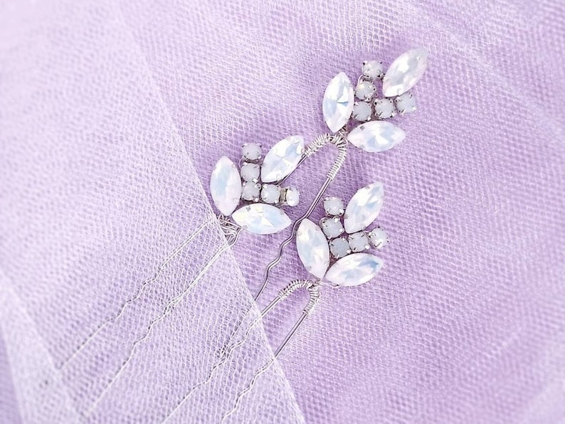 May include: Three silver hair clips with white crystal embellishments. The clips are arranged in a cluster on a light purple background.