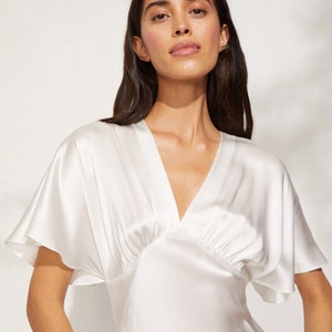 May include: A white silk dress with a V-neckline and short, wide sleeves. The dress has a gathered detail at the bust and a flowing skirt.