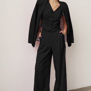 Pode incluir: Um blazer preto com um colete preto e cal&ccedil;as pretas de perna larga. A pessoa est&aacute; usando &oacute;culos de sol pretos.