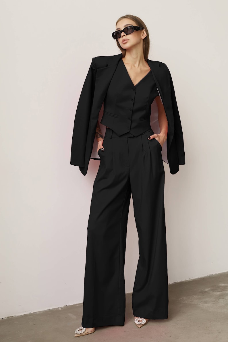 Pode incluir: Um blazer preto com um colete preto e cal&ccedil;as pretas de perna larga. A pessoa est&aacute; usando &oacute;culos de sol pretos.