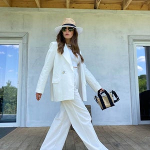 May include: A woman wearing a white pantsuit with a wide-brimmed straw hat and sunglasses. She is carrying a clear plastic bag with a black strap.