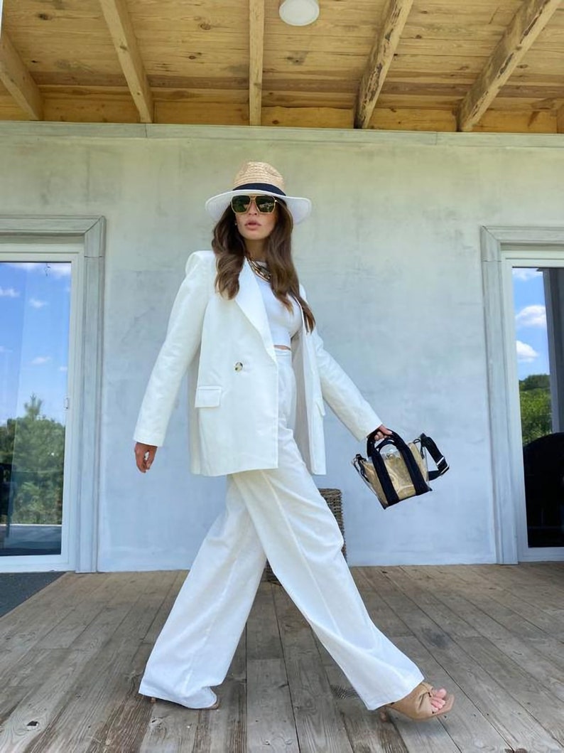May include: A woman wearing a white pantsuit with a wide-brimmed straw hat and sunglasses. She is carrying a clear plastic bag with a black strap.