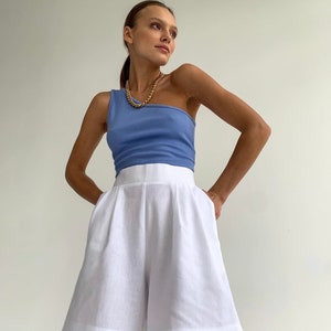 Pode incluir: Uma mulher vestindo um top azul claro de um ombro s&oacute; e shorts de linho branco. Ela est&aacute; usando um colar de corrente de ouro.