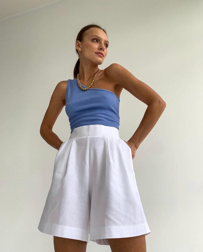 Pode incluir: Uma mulher vestindo um top azul claro de um ombro s&oacute; e shorts de linho branco. Ela est&aacute; usando um colar de corrente de ouro.