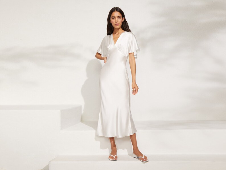 May include: A white satin midi dress with a V-neckline and short, flared sleeves. The dress has a fitted bodice and a flowing skirt.