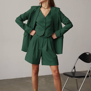 May include: A woman wearing a green blazer, waistcoat, and shorts with a gold chain necklace and lime green high-heeled sandals.