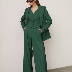 May include: A woman wearing a dark green suit with a blazer, waistcoat, and wide-leg trousers. She is wearing black sunglasses and has her hands in her pockets.