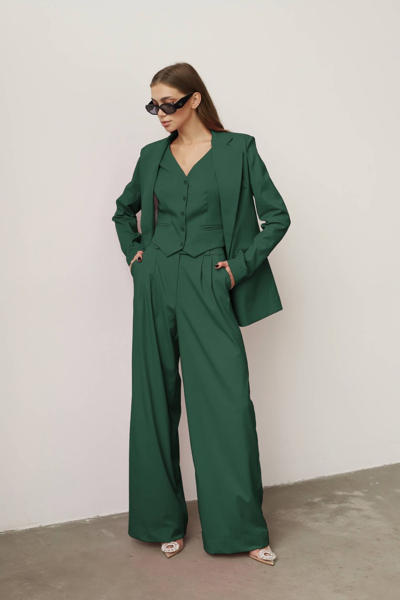 May include: A woman wearing a dark green suit with a blazer, waistcoat, and wide-leg trousers. She is wearing black sunglasses and has her hands in her pockets.