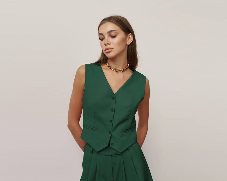 May include: A dark green sleeveless waistcoat with a button fastening and a gold chain necklace. The waistcoat has a v-neck and a tailored fit.