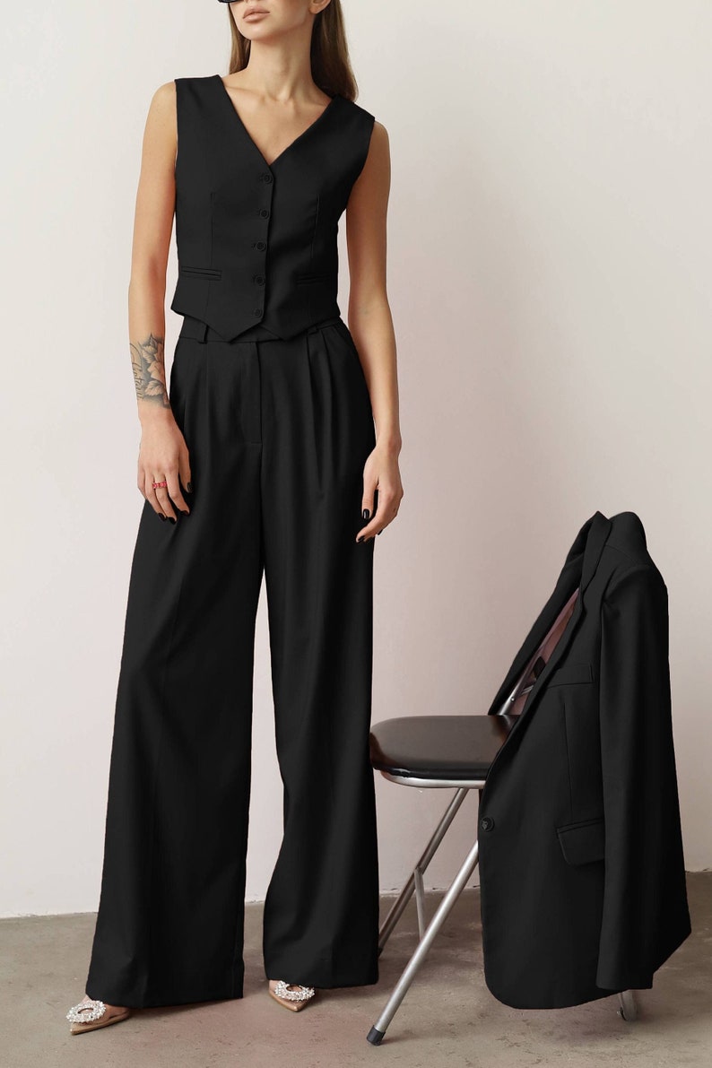 Pode incluir: Um colete preto sem mangas com bot&otilde;es, cal&ccedil;as pretas de perna larga e um blazer preto. O visual &eacute; complementado por um par de sapatos adornados com prata e branco.