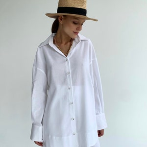 Pode incluir: Uma camisa branca de linho com mangas compridas e fecho de bot&otilde;es. A camisa tem um corte descontra&iacute;do e uma silhueta ligeiramente oversized. A modelo est&aacute; a usar um chap&eacute;u de palha com uma faixa preta.