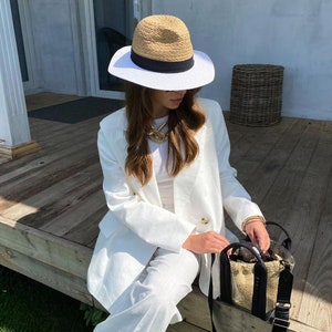 May include: A woman wearing a white linen blazer and pants with a wide-brimmed straw hat with a black band. She is holding a clear tote bag with a black strap and a woven straw base.