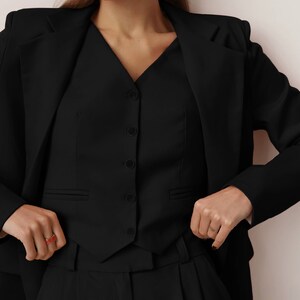 Pode incluir: Um blazer preto com um colete preto por baixo. O colete tem um decote em V e cinco bot&otilde;es. A pessoa est&aacute; usando cal&ccedil;as pretas.