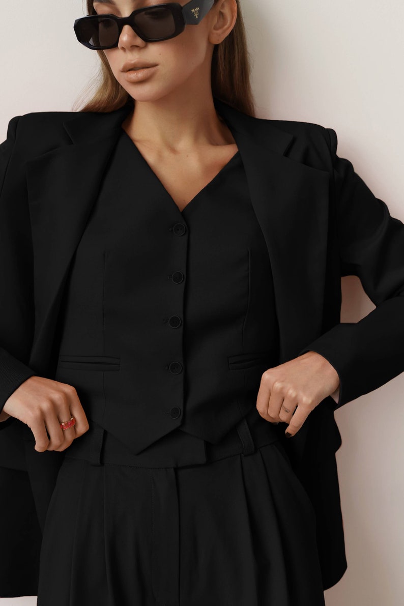 Pode incluir: Um blazer preto com um colete preto por baixo. O colete tem um decote em V e cinco bot&otilde;es. A pessoa est&aacute; usando cal&ccedil;as pretas.
