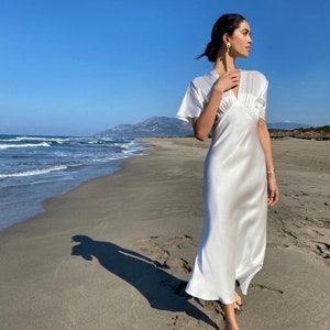 May include: A woman wearing a white satin dress with a V-neckline and short sleeves stands on a sandy beach with the ocean in the background.