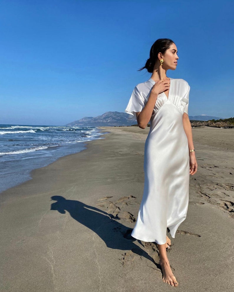 May include: A woman wearing a white satin dress with a V-neckline and short sleeves stands on a sandy beach with the ocean in the background.