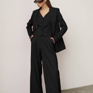 Pode incluir: Um blazer preto com um colete preto e cal&ccedil;as pretas de perna larga. A pessoa est&aacute; usando &oacute;culos de sol pretos.