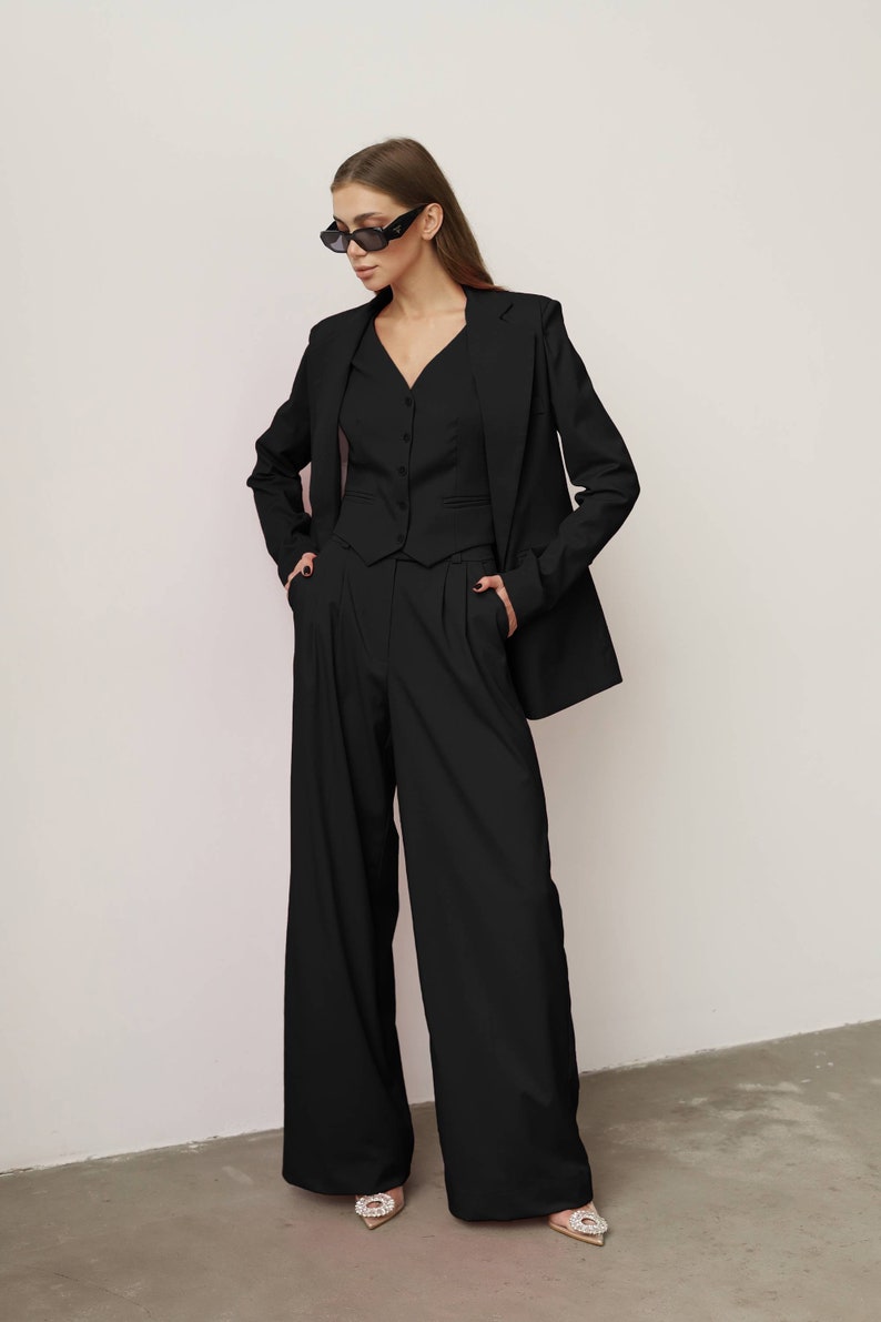 Pode incluir: Um blazer preto com um colete preto e cal&ccedil;as pretas de perna larga. A pessoa est&aacute; usando &oacute;culos de sol pretos.