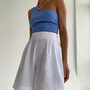 Pode incluir: Uma mulher est&aacute; vestindo um top azul claro de um ombro s&oacute; e shorts brancos de cintura alta. Ela est&aacute; usando um colar de corrente de ouro.