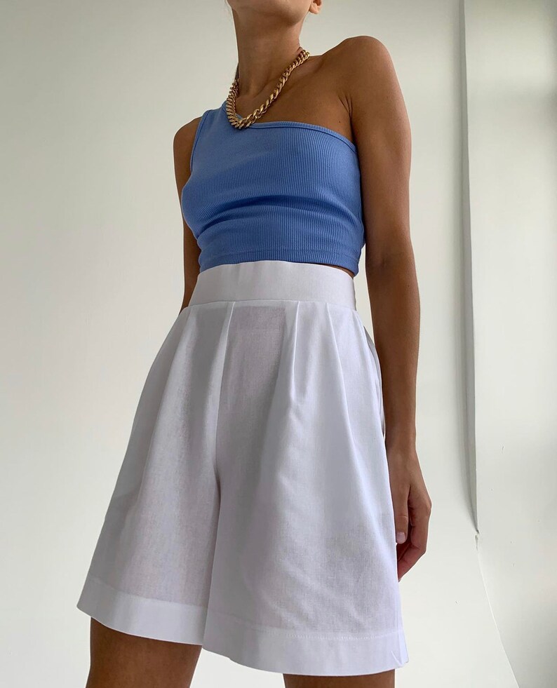 Pode incluir: Uma mulher est&aacute; vestindo um top azul claro de um ombro s&oacute; e shorts brancos de cintura alta. Ela est&aacute; usando um colar de corrente de ouro.