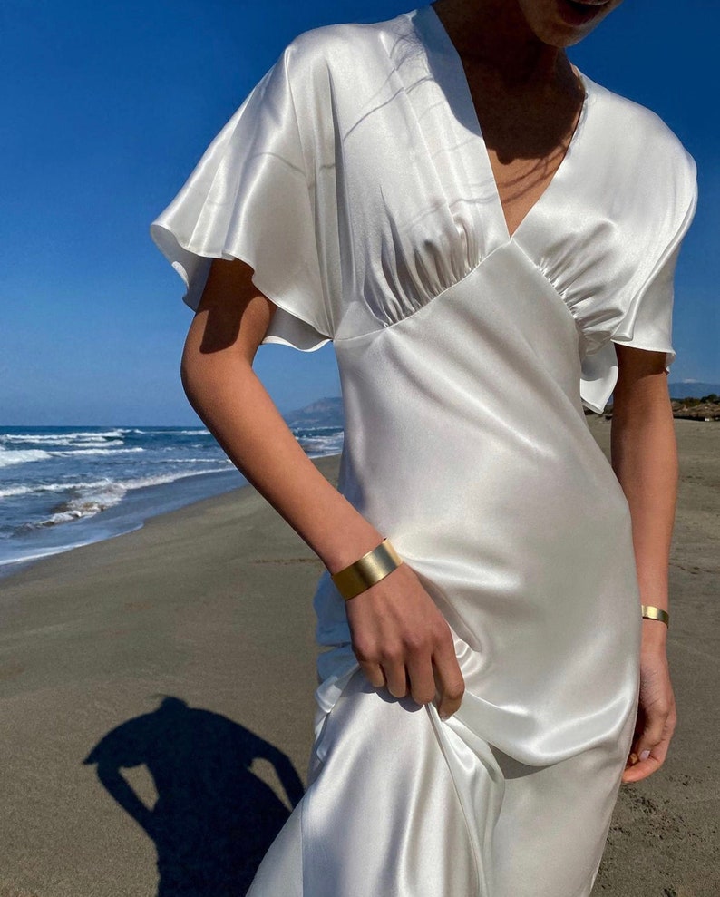 May include: A white satin dress with a v-neckline and short sleeves. The dress has a flowing silhouette and is perfect for a summer wedding or special occasion.