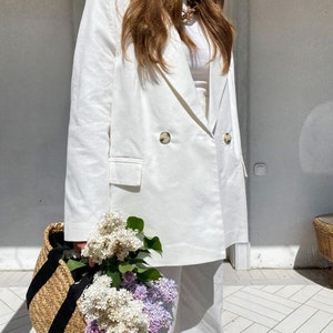 May include: A white linen blazer with a double-breasted closure and a single button. The blazer is worn with white pants and a white t-shirt. The person is holding a bouquet of white and purple flowers in a woven basket with black straps.