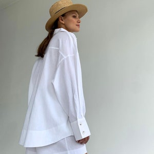 Pode incluir: Uma camisa branca de linho de manga comprida e caimento solto, combinada com shorts de linho branco combinando. A modelo est&aacute; usando um chap&eacute;u de palha.