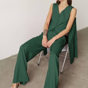 May include: A woman wearing a dark green pantsuit with a sleeveless vest and a blazer. She is sitting on a metal chair and wearing clear heels with rhinestone embellishments.