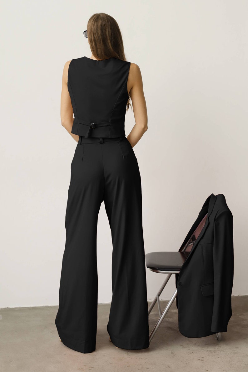 Pode incluir: Um colete preto sem mangas com cinto e cal&ccedil;as largas. Um blazer preto est&aacute; drapeado sobre uma cadeira.