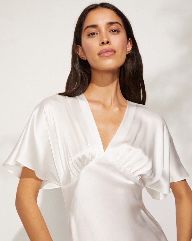 May include: A white silk dress with a V-neckline and short, wide sleeves. The dress has a gathered detail at the bust and a flowing skirt.