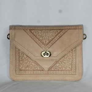 Könnte beinhalten: Beige Leder-Umhängetasche mit geprägtem floralem Design. Die Tasche hat einen Klappenverschluss mit einem messingfarbenen Verschluss und Metallringen für einen Riemen. Die Tasche ist rechteckig mit dekorativem Zierstich.