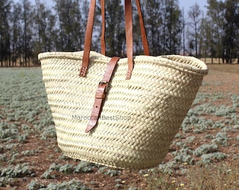 May include: A woven straw tote bag with brown leather handles and a buckle closure. The bag is rectangular with a wide opening and is held against a field of green plants and trees.