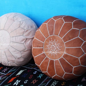 May include: Two round leather poufs. One is light beige, the other is brown. Both have a starburst design in the center and are stitched with white thread. The poufs are on a patterned rug.