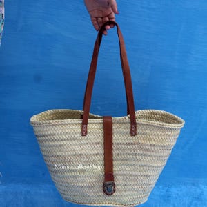 May include: A woven straw tote bag with brown leather handles and accents. The bag has a wide opening and a rectangular base. The handles are attached with decorative stitching. The bag is held against a blue background. The text "MoroccanBestShop" is visible on the bag.